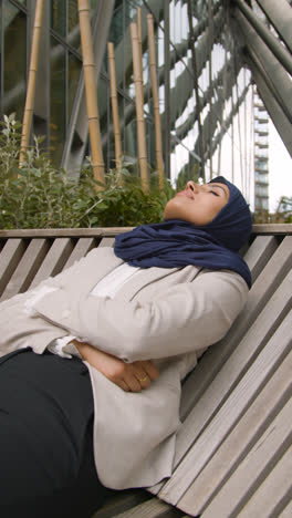 Vertical-Video-Of-Muslim-Businesswoman-Resting-On-Bench-Outdoors-In-City-Gardens-Or-Park